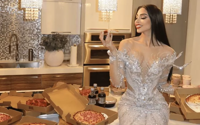 Daniela Reyes posando con un vestido de gala de cristales en una cocina mientras sostiene un trozo de pizza.